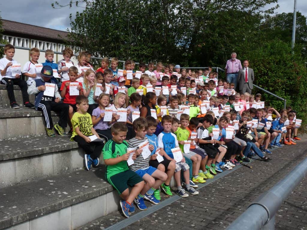 Eine große Gruppe Kinder sitzt auf einer Außentreppe, hält Zertifikate in den Händen und lächelt in die Kamera. Oben rechts stehen zwei Erwachsene und beaufsichtigen die Gruppe. Im Hintergrund sind Bäume und ein Gebäude zu sehen.