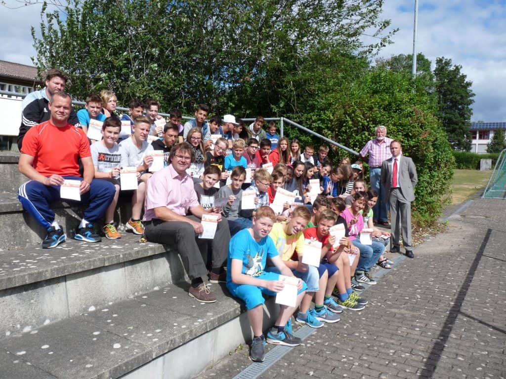 Eine große Gruppe Kinder und einige Erwachsene sitzen auf den Außentreppen, viele halten Zertifikate in den Händen. Einige Erwachsene stehen in der Nähe. Es ist ein sonniger Tag mit viel Grün und Gebäuden im Hintergrund.