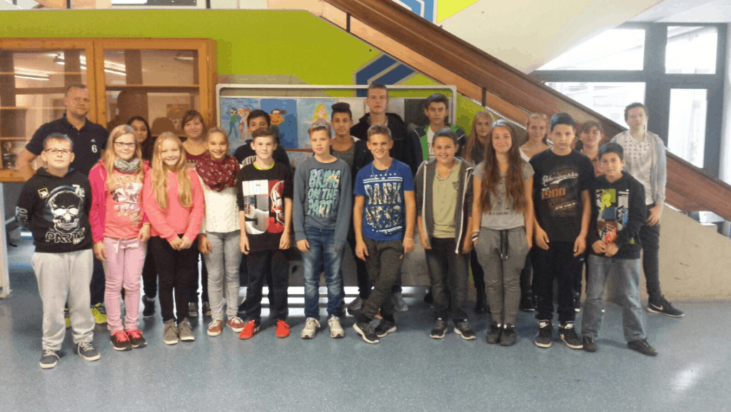 Eine Gruppe von Schülern und ein paar Erwachsenen stehen zusammen in einem Raum vor einer bunten Wand und Treppe und lächeln in die Kamera. Die Umgebung scheint eine Schule zu sein.