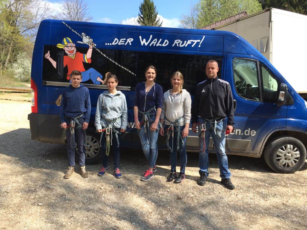 Fünf Personen mit Klettergurten stehen an einem sonnigen Tag im Freien vor einem blauen Lieferwagen mit der Aufschrift „DER WALD RUFT!“ und einem Cartoon-Kletterer an der Seite.