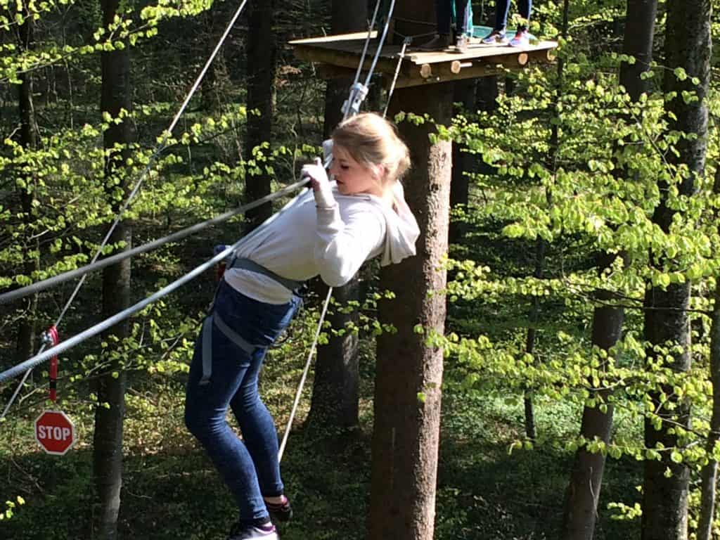 Ein Mädchen in weißem Kapuzenpullover und Jeans balanciert auf Seilen in einem Walderlebnispark. Sie klammert sich an die Seile, lehnt sich zurück und wirkt konzentriert. Hinter ihr ist eine Plattform zu sehen, und Bäume umgeben das Gelände.