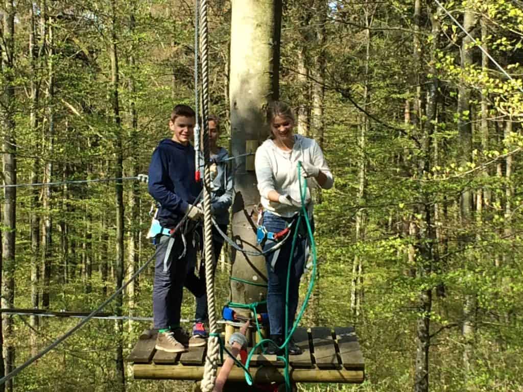 Drei Personen mit Klettergurten stehen auf einer Holzplattform, die an einem Baum im Wald befestigt ist, und bereiten sich auf einen Hochseilgarten oder eine Seilrutsche vor. Grünes Laub umgibt sie.