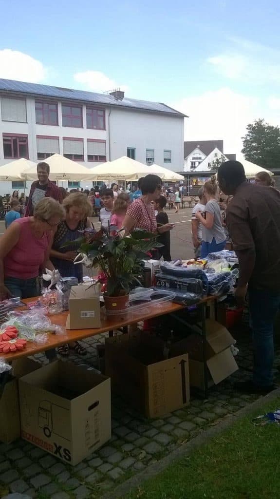 Menschen stöbern auf einem Markt oder Jahrmarkt an Tischen. Kisten und verschiedene Waren sind ausgestellt. Andere Menschen sitzen unter weißen Sonnenschirmen im Hintergrund in der Nähe eines großen Gebäudes. Der Himmel ist teilweise bewölkt.