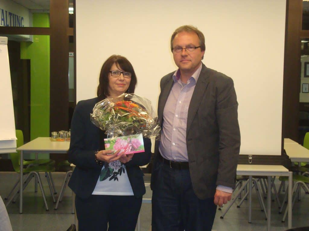 Eine Frau mit einem Blumenstrauß und einer Karte steht neben einem Mann in Geschäftskleidung. Sie posieren in einem Innenbereich vor einer Projektionsfläche und Tischen.