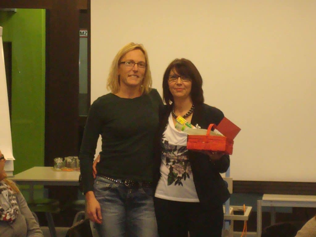 Zwei Frauen stehen nebeneinander, eine hat den Arm um die andere gelegt. Die rechte Frau hält einen roten Korb mit verschiedenen Gegenständen. Sie befinden sich in einem Raum mit einem Whiteboard und einer grünen Wand im Hintergrund.