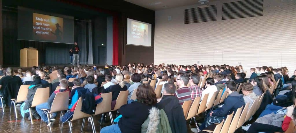 Eine große Gruppe von Menschen sitzt in Stuhlreihen vor einer Bühne, auf der ein Redner einen Vortrag hält. Auf einem Bildschirm erscheint der deutsche Text: „Sieh auf, geh raus und machs einfach!“ Der Raum ist gut beleuchtet, mit Holzböden und hohen Decken.