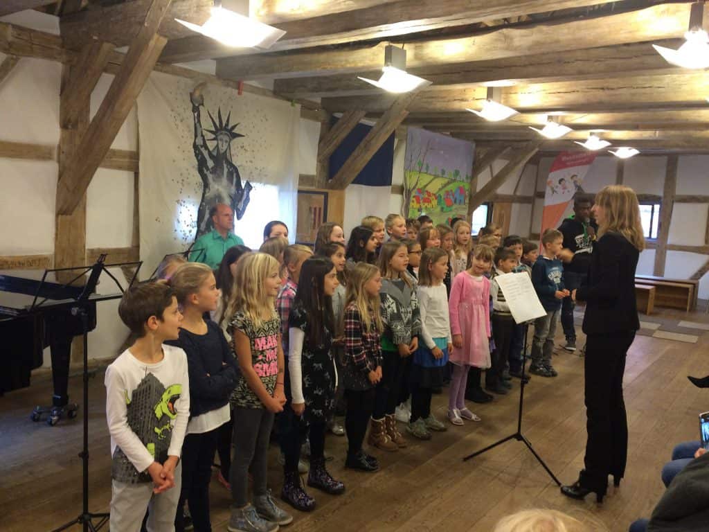 Eine Gruppe Kinder steht in Reihen in einem rustikalen Raum mit Holzbalken und singt. Eine erwachsene Frau dirigiert sie, in der Nähe stehen Musiker. Im Hintergrund hängen bunte Banner an den Wänden.