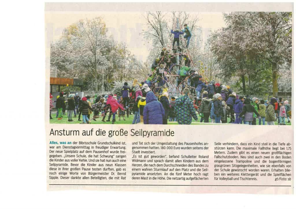 Kinder in bunter Winterkleidung klettern und spielen auf einer großen Seilpyramide auf einem verschneiten Schulhof, während Erwachsene daneben stehen und zuschauen. Im Hintergrund sind Bäume und Spielgeräte zu sehen.