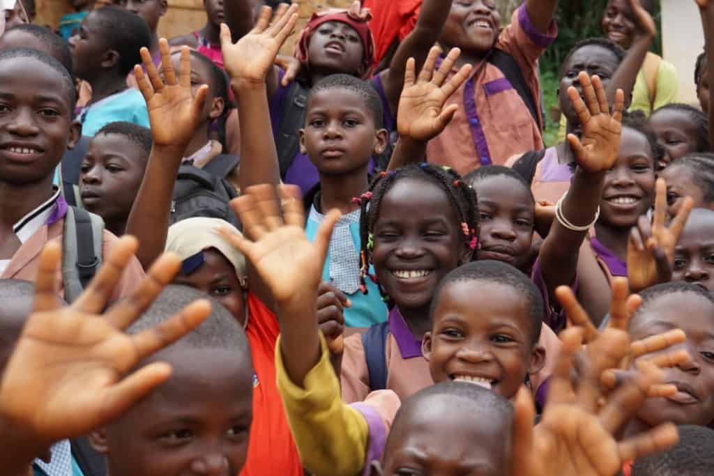 Eine Gruppe lächelnder Kinder in Schuluniformen winkt begeistert in die Kamera und zeigt Aufregung und Freude. Einige tragen Rucksäcke, und die Kinder stehen dicht gedrängt im Freien.