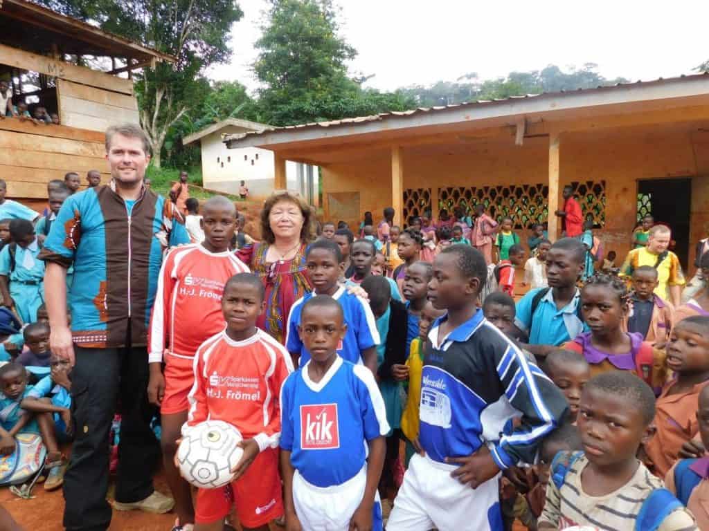 Eine Gruppe Kinder in Fußballtrikots steht mit zwei Erwachsenen im Freien, umgeben von weiteren Kindern und einem gelben Gebäude im Hintergrund. Ein Kind hält einen Fußball. Hinter dem Gebäude sind Bäume und Grünflächen zu sehen.