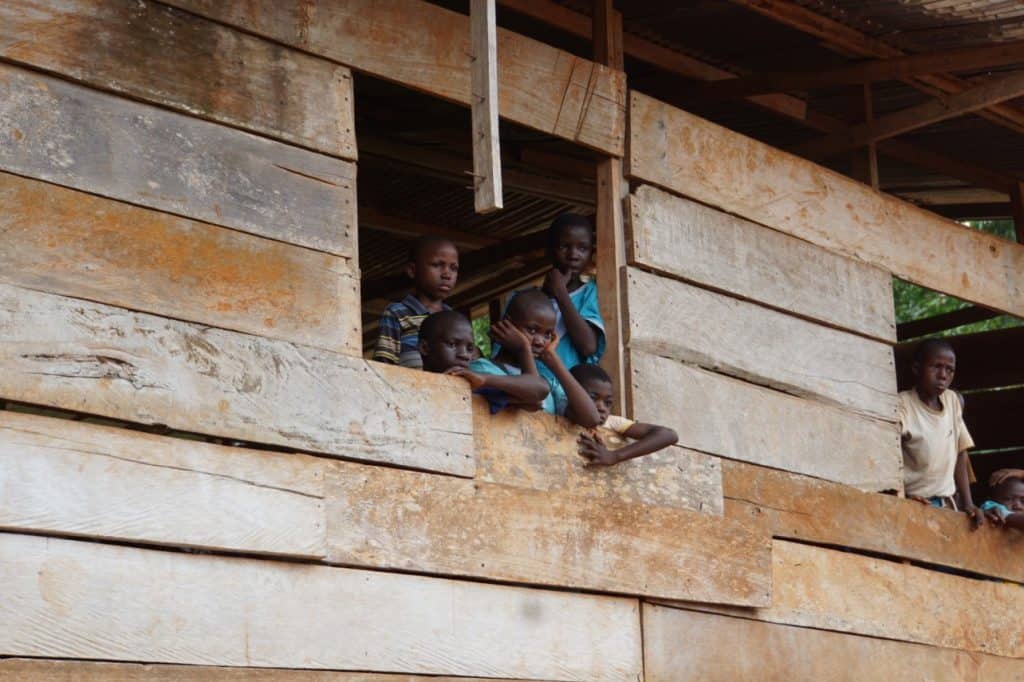 Mehrere Kinder lehnen aus einem großen Fenster in einem Holzgebäude und schauen mit neugierigen und nachdenklichen Blicken nach draußen. Das Gebäude ist rustikal und besteht aus verwitterten Holzbrettern.