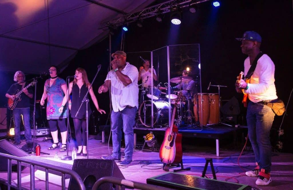 Eine Band spielt unter violettem Licht auf der Bühne. Drei Sänger stehen vorne, einer hält ein Mikrofon. Im Hintergrund spielen Musiker Gitarre, Schlagzeug und Congas. Verschiedene Instrumente und Bühnentechnik sind zu sehen.