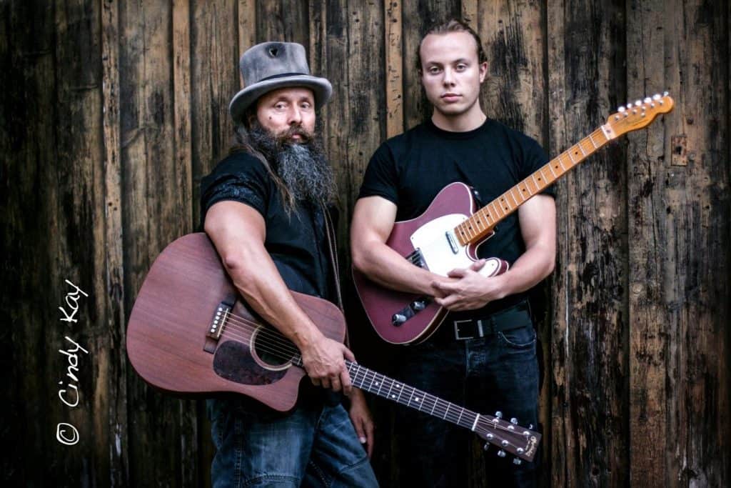 Vor einer rustikalen Holzwand stehen zwei Männer, jeder hält eine Gitarre. Der linke Mann hat einen langen Bart, trägt einen Hut und hält eine Akustikgitarre, der rechte eine E-Gitarre.