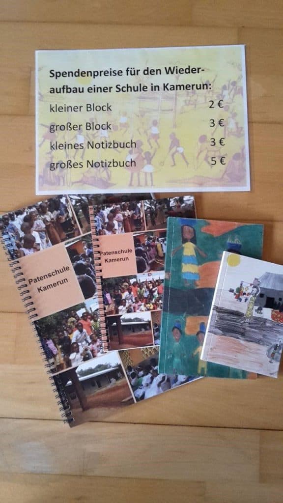 Fünf Notizbücher mit bunten Einbänden und Fotos liegen auf einem Holzboden. Darüber hängt ein Schild mit den Spendenpreisen für den Wiederaufbau einer Schule in Kamerun in deutscher Sprache, mit Preisen für verschiedene Notizheftgrößen.