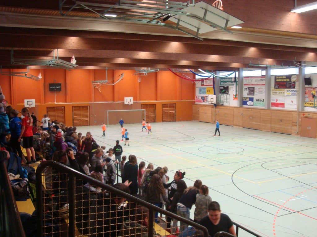 Eine Menschenmenge verfolgt ein Jugend-Hallenfußballspiel in einer Turnhalle. Mehrere Kinder in bunten Trikots spielen auf dem Platz, während Zuschauer auf Tribünen an einer Seite des Raumes sitzen. An den Wänden hängen Banner und Basketballkörbe.