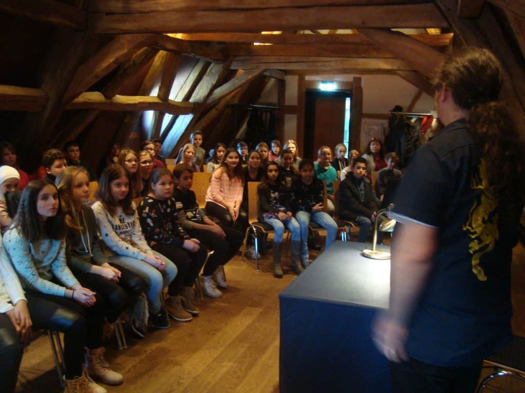 Eine Gruppe Kinder sitzt aufmerksam in einem Raum mit Holzbalken und blickt auf einen Erwachsenen, der vorne an einem Tisch steht, was an ein Klassenzimmer oder eine pädagogische Präsentationsumgebung erinnert.