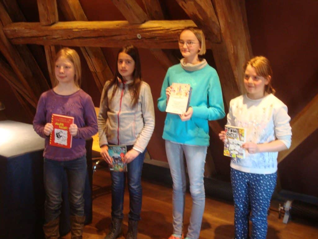 Vier Mädchen stehen drinnen vor Holzbalken, halten jeweils ein Buch in der Hand und blicken in die Kamera. Sie sind leger gekleidet und scheinen an einer Buchveranstaltung oder einem Wettbewerb teilzunehmen.