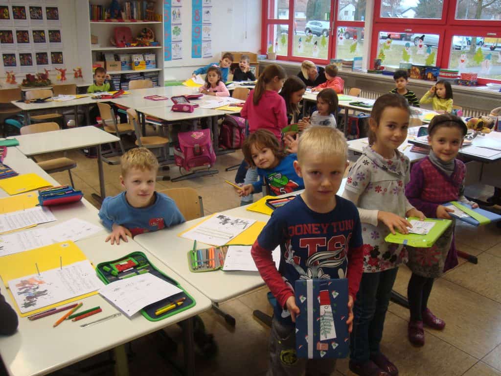 Ein Klassenzimmer voller kleiner Kinder, die an ihren Tischen sitzen und an ihren Aufgaben arbeiten. Einige Schüler stehen vorne, halten bunte Gegenstände in den Händen und lächeln in die Kamera. Der Raum ist hell, große Fenster und verschiedene Schulmaterialien sind sichtbar.