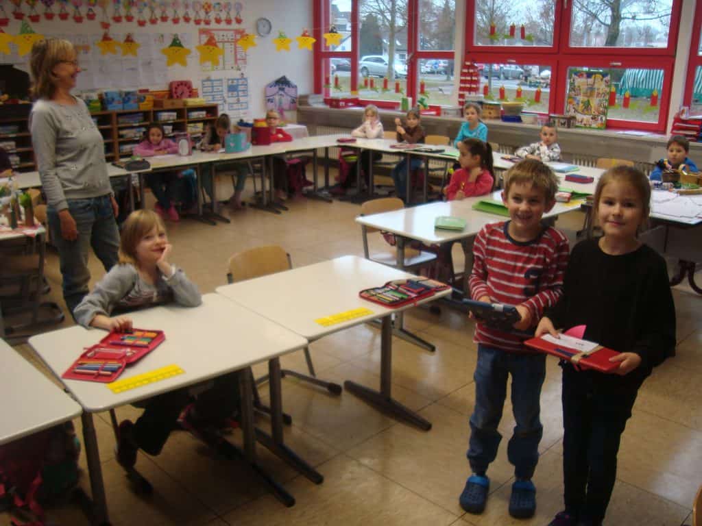 Ein Klassenzimmer mit kleinen Kindern, die an ihren Tischen sitzen und teilweise in die Kamera blicken. Zwei Kinder stehen vorne und halten Schulsachen in den Händen, während links eine Lehrerin steht. Der Raum ist mit farbenfrohen Kunstwerken und Sternen dekoriert.