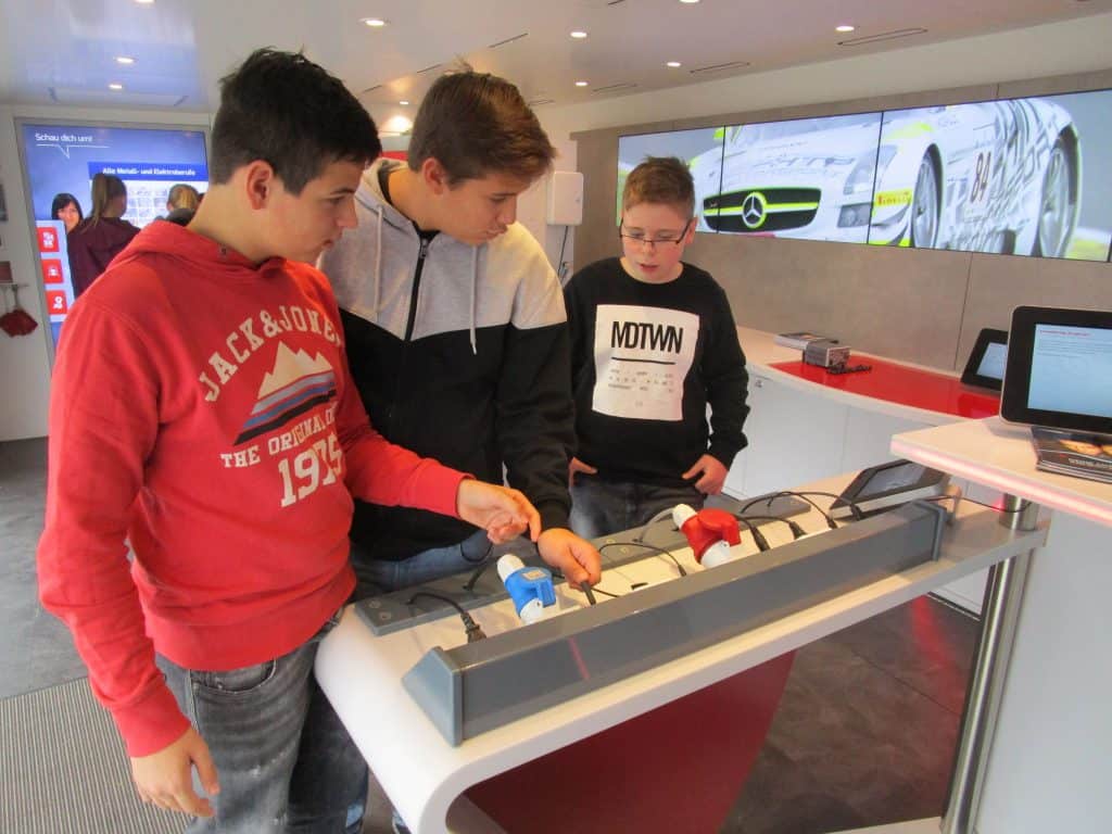 Drei Jungen stehen an einem interaktiven Ausstellungstisch und untersuchen kleine Modelle mit Knöpfen und Bildschirmen in einem modernen, gut beleuchteten Raum mit einem Autobild an der Wand im Hintergrund.