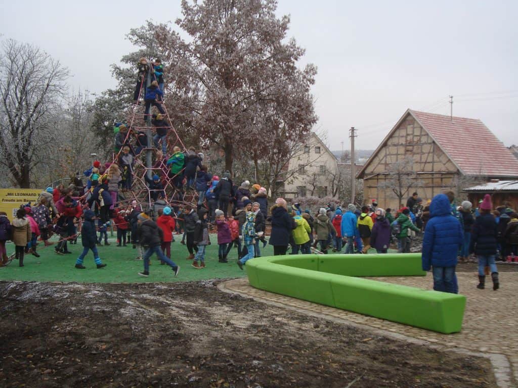 Kinder spielen auf einem Spielplatz mit einem Kletterseil, während sich andere in der Nähe versammeln. Der Spielplatz hat einen grünen Boden und im Hintergrund sind an einem bewölkten Tag Bäume und Gebäude zu sehen.