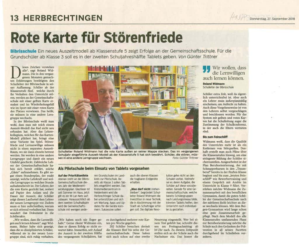 Ein Mann im Anzug sitzt an einem Schreibtisch in einem Klassenzimmer, hält eine rote Karte hoch und lächelt in die Kamera. Auf dem Schreibtisch und den Regalen im Hintergrund sind Bücher, Papiere und bunte Schulmaterialien zu sehen.