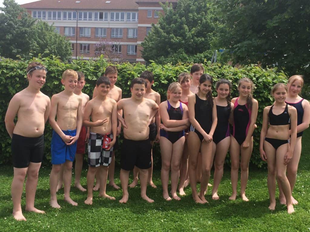 Eine Gruppe Kinder in Badeanzügen steht barfuß im Gras, lächelt und posiert für ein Foto im Freien mit Büschen und einem Schulgebäude im Hintergrund.