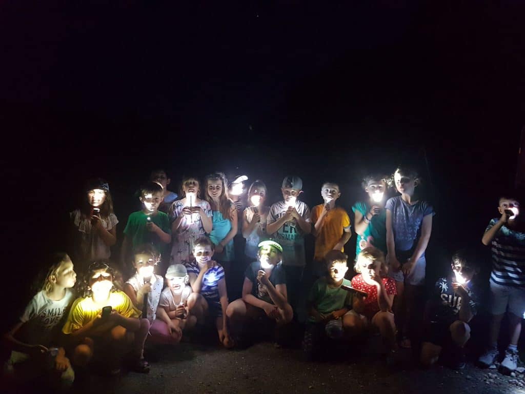 Eine Gruppe von Kindern sitzt und steht dicht beieinander im Dunkeln und leuchtet sich mit Taschenlampen ins Gesicht, wodurch ein unheimliches Leuchten entsteht. Der Hintergrund ist fast komplett schwarz.