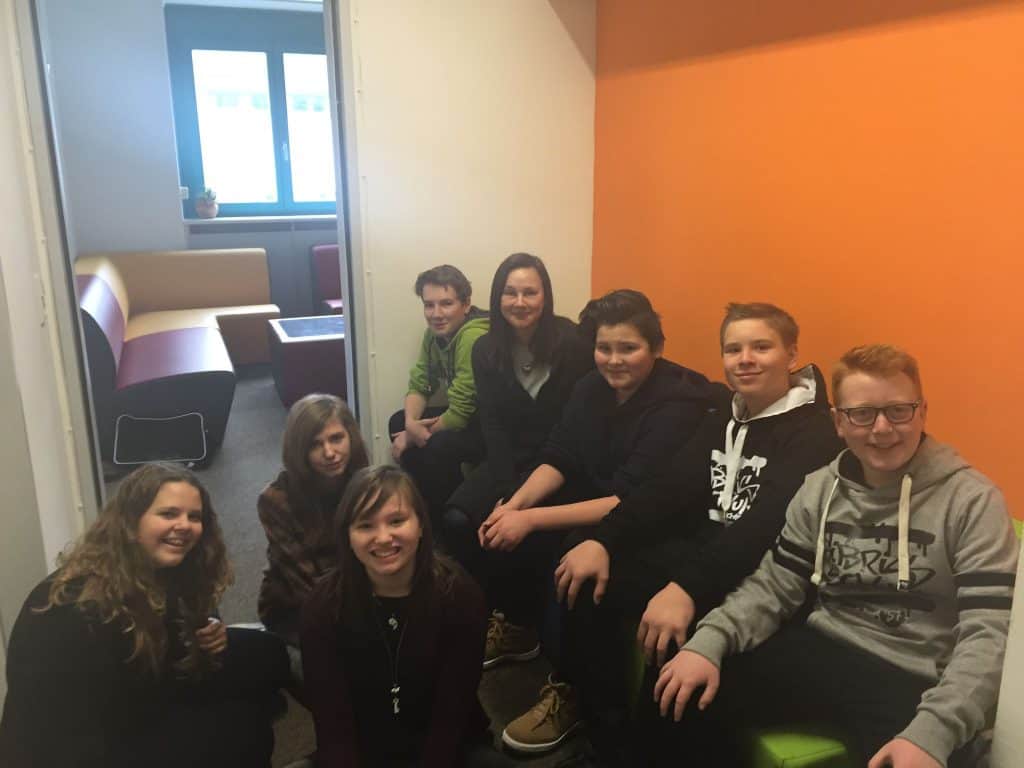 Eine Gruppe von sieben Teenagern und einem Erwachsenen sitzt lächelnd in einem Raum mit orangefarbenen und weißen Wänden, in der Nähe einer offenen Tür, die zu einem anderen Raum mit Sofas und einem Fenster führt.