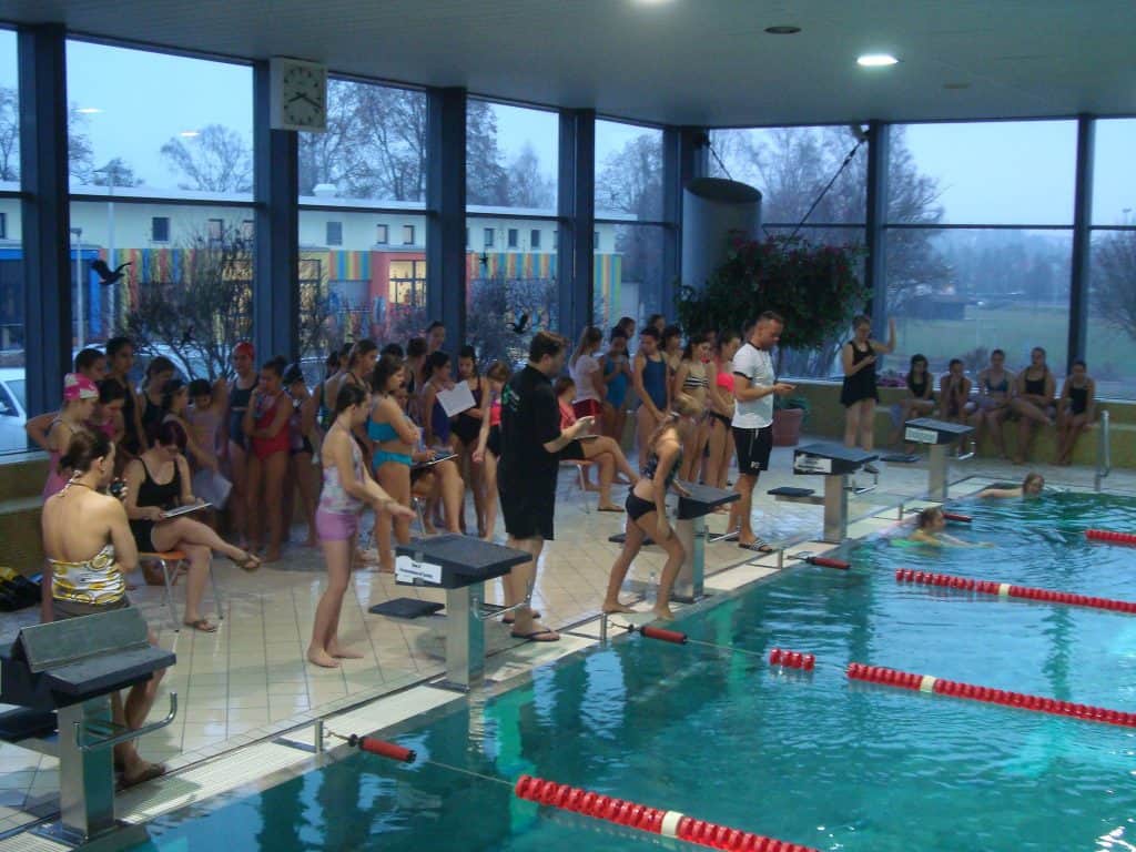 Eine Gruppe Schwimmer in Badeanzügen steht am Rand eines Hallenbads und bereitet sich auf einen Wettkampf vor. Trainer und Offizielle sind anwesend und der Beckenbereich verfügt über große Fenster mit Blick ins Freie.