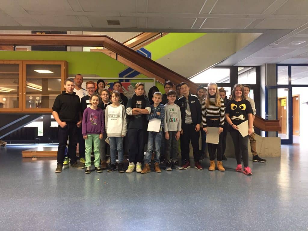 Eine Gruppe von Schülern und Erwachsenen steht gemeinsam vor einer Treppe in einem Gebäude und posiert für ein Gruppenfoto. Die Szenerie scheint ein Schulflur mit hellen Wänden und Fenstern im Hintergrund zu sein.