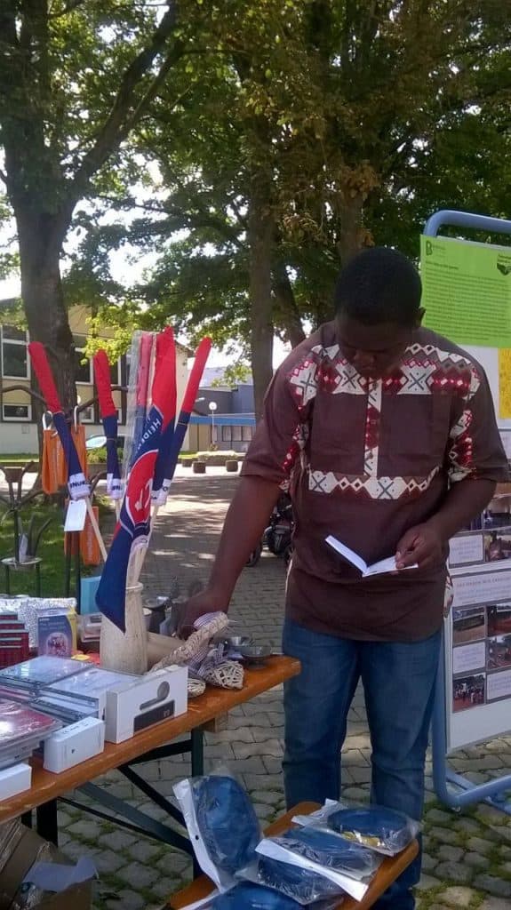 Ein Mann steht im Freien an einem Tisch mit Büchern, Kunsthandwerk und Flaggen und liest eine Broschüre. Hinter ihm stehen Bäume, Schautafeln mit Informationen und weitere Tische auf einem Kopfsteinpflasterplatz.