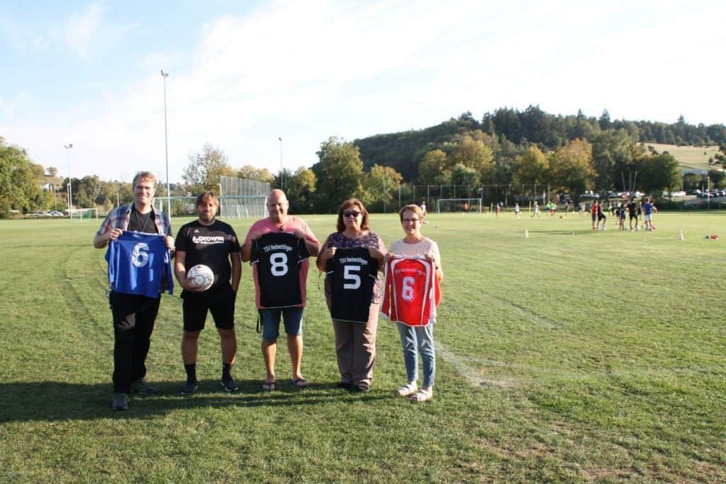 Fünf Personen stehen mit Fußballtrikots und einem Ball in den Händen auf einem Rasensportplatz. Im Hintergrund sind Bäume und Hügel zu sehen. Drei tragen Sonnenbrillen, und in der Ferne spielt eine Gruppe.