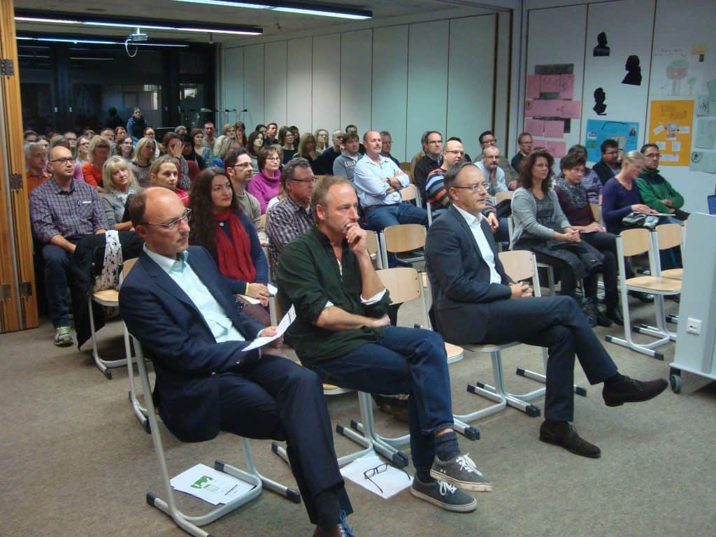 Eine Gruppe Erwachsener sitzt aufmerksam in Stuhlreihen in einem Klassen- oder Konferenzraum und hört einem Vortrag zu. Vier Personen sitzen in der ersten Reihe, viele weitere dahinter. An den Wänden hängen bunte Poster.