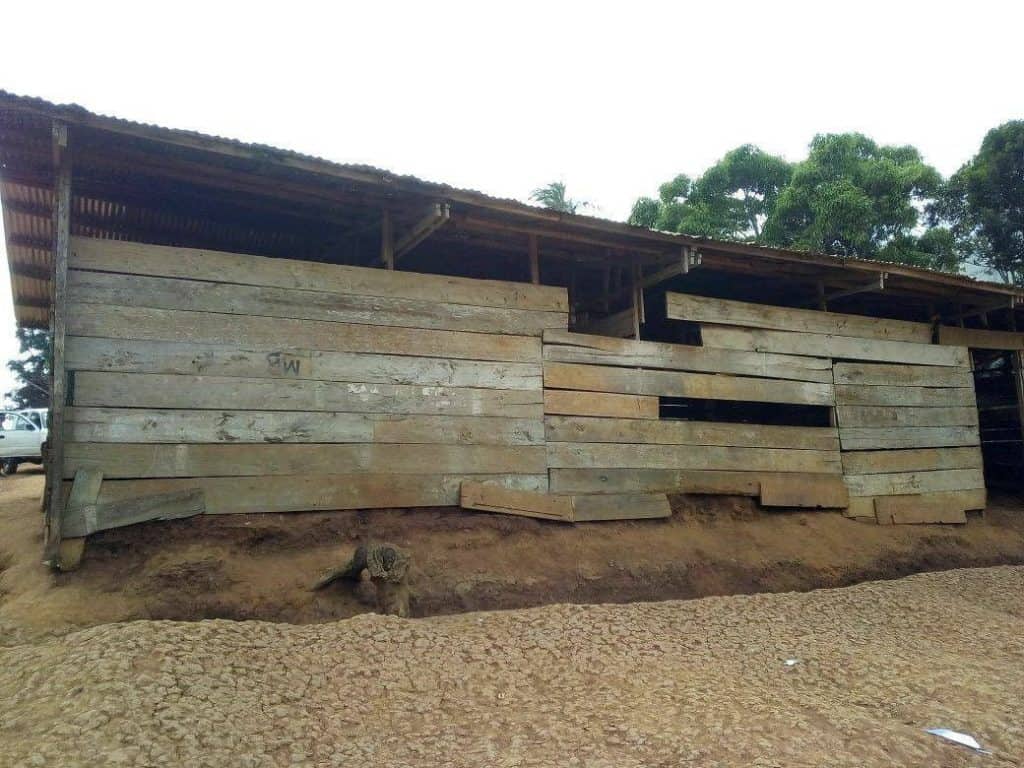 Auf einem unbefestigten Fleck steht ein rustikales Holzgebäude mit unebenen Brettern und Lücken in den Wänden. Das Fundament ist leicht erhöht, und das Dach ist aus Wellblech. Im Hintergrund sind Bäume und ein bewölkter Himmel zu sehen.