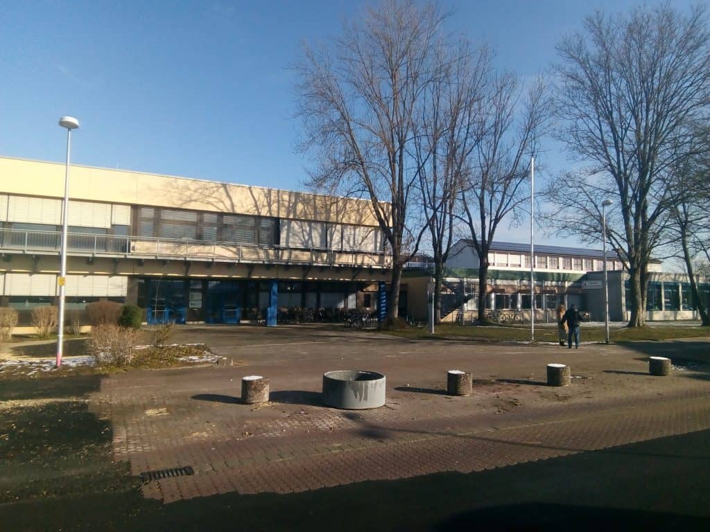 Ein zweistöckiges Schulgebäude mit beigen und blauen Wänden steht hinter einem gepflasterten Innenhof mit Bäumen, Laternenpfählen und einem runden Betonkübel. Zwei Menschen gehen spazieren; auf dem Gras sind Schneeflecken zu sehen.