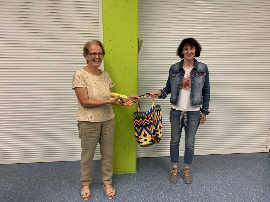 Zwei Frauen stehen drinnen vor einem weißen Fensterladen und einer grünen Säule. Eine Frau reicht der anderen einen gelben Gegenstand und eine gemusterte Tasche. Beide Frauen lächeln und sind leger gekleidet.