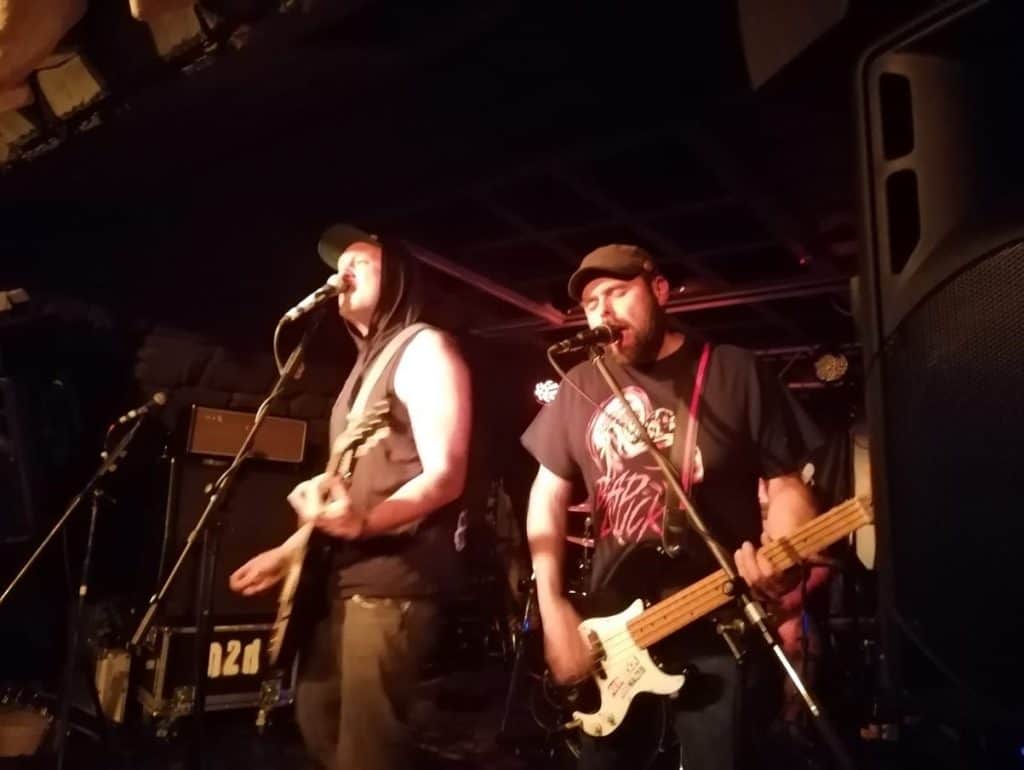 Zwei Musiker treten bei gedämpftem Licht auf der Bühne auf; beide singen in Mikrofone und spielen E-Gitarre. Einer trägt einen ärmellosen Hoodie und eine Mütze, der andere ein T-Shirt und eine Mütze. Im Hintergrund ist Musikinstrument zu sehen.