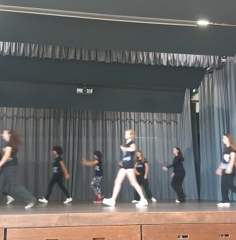 Eine Gruppe von schwarz-weiß gekleideten Jugendlichen übt auf einer Bühne mit grauen Vorhängen im Hintergrund eine Tanznummer ein. Einige der Tänzerinnen und Tänzer befinden sich mitten im Schritt, was ein Gefühl der Bewegung vermittelt.