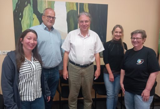 Fünf Erwachsene stehen in einem Haus und lächeln in die Kamera. An der Wand hinter ihnen ist ein Gemälde mit Bäumen zu sehen. Die Gruppe wirkt entspannt und freundlich; drei Männer und zwei Frauen sind zu sehen.