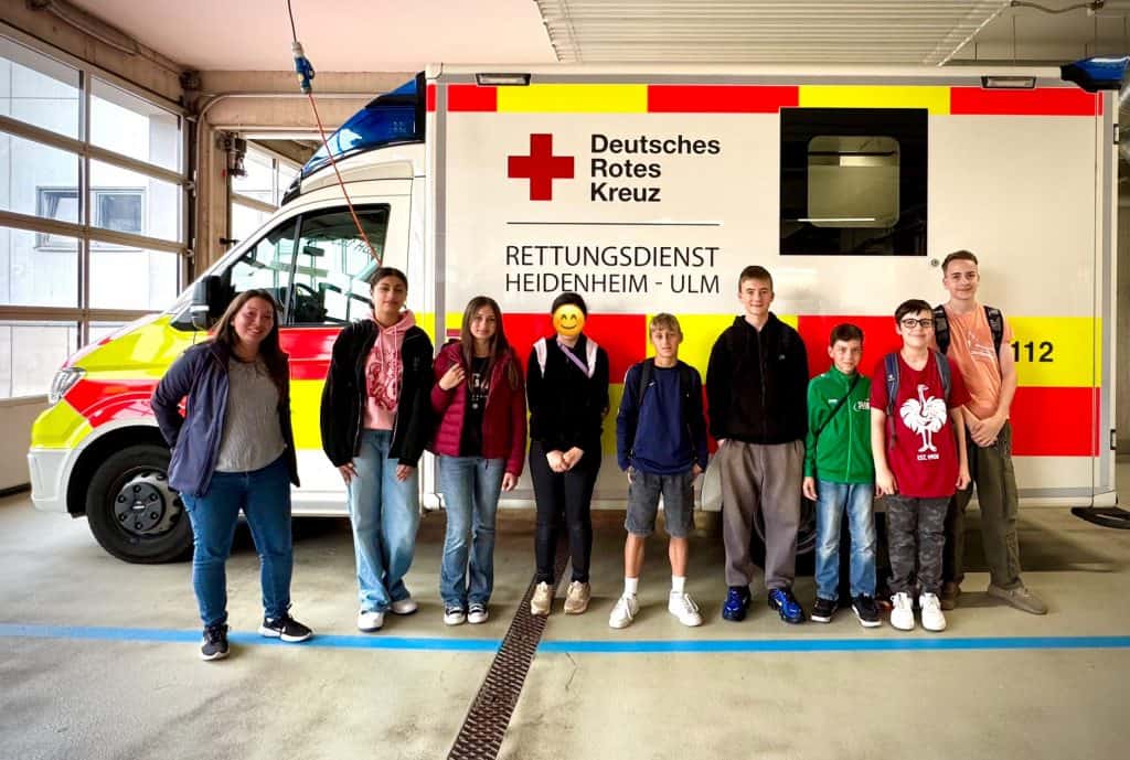 Eine Gruppe von acht jungen Menschen steht vor einem Rettungswagen des Deutschen Roten Kreuzes in einer Garage. Der Krankenwagen ist weiß mit roten und gelben Markierungen und deutschem Text. Die Gruppe ist leger gekleidet und lächelt in die Kamera.