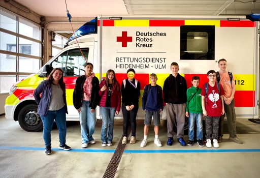 Eine Gruppe von acht jungen Menschen steht vor einem Rettungswagen des Deutschen Roten Kreuzes in einer Garage. Der Krankenwagen ist weiß mit roten und gelben Markierungen und deutschem Text. Die Gruppe ist leger gekleidet und lächelt in die Kamera.