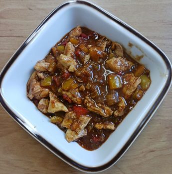 Eine quadratische weiße Schale, gefüllt mit einem stückigen Eintopf mit Hühnerfleischstücken, gewürfelter roter und gelber Paprika und einer reichhaltigen braunen Soße, die auf einer hellen Holzunterlage steht.