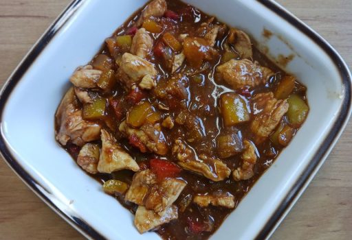 Eine quadratische weiße Schale, gefüllt mit einem stückigen Eintopf mit Hühnerfleischstücken, gewürfelter roter und gelber Paprika und einer reichhaltigen braunen Soße, die auf einer hellen Holzunterlage steht.
