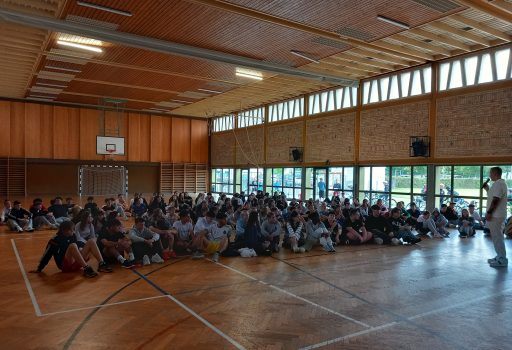 Eine große Gruppe von Schülern sitzt auf dem Boden einer geräumigen Turnhalle mit Holzböden und hohen Fenstern und blickt aufmerksam auf einen Erwachsenen, der vorne steht.