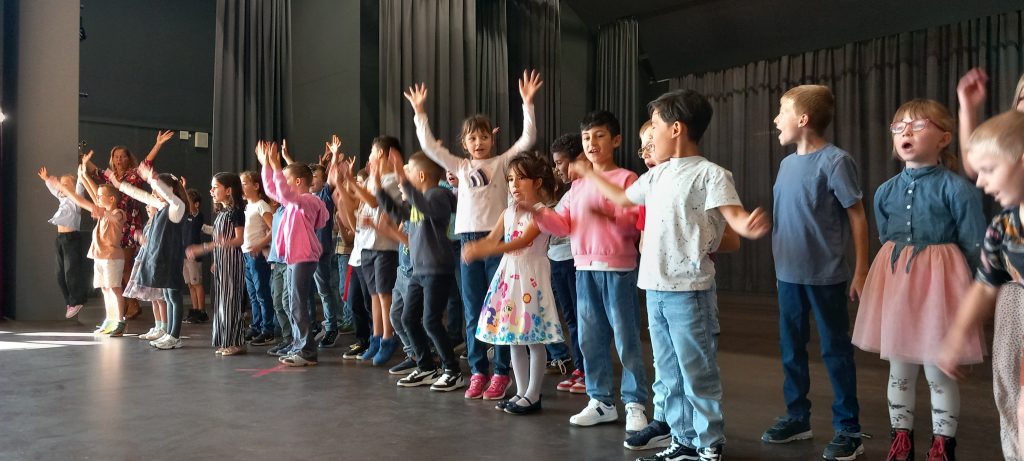 Eine Gruppe kleiner Kinder steht in einer Reihe auf einer Bühne, einige mit erhobenen Armen und lächelnd, als ob sie singen oder etwas vorführen würden. Hinter ihnen hängen dunkle Vorhänge, und von links scheint das Sonnenlicht herein.