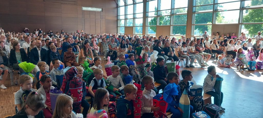 Eine große Gruppe von Kindern und Erwachsenen sitzt in einer sonnenbeschienenen Halle mit Holzwänden und großen Fenstern. Die Kinder, von denen viele bunte, kegelförmige Geschenke in der Hand halten, sitzen vorne; die Erwachsenen füllen die Sitzreihen hinter ihnen.