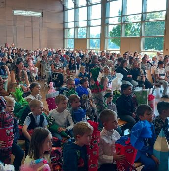 Eine große Gruppe von Kindern und Erwachsenen sitzt in einer sonnenbeschienenen Halle mit Holzwänden und großen Fenstern. Die Kinder, von denen viele bunte, kegelförmige Geschenke in der Hand halten, sitzen vorne; die Erwachsenen füllen die Sitzreihen hinter ihnen.