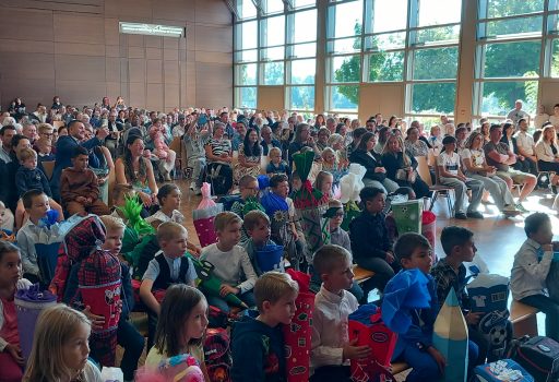 Eine große Gruppe von Kindern und Erwachsenen sitzt in einer sonnenbeschienenen Halle mit Holzwänden und großen Fenstern. Die Kinder, von denen viele bunte, kegelförmige Geschenke in der Hand halten, sitzen vorne; die Erwachsenen füllen die Sitzreihen hinter ihnen.