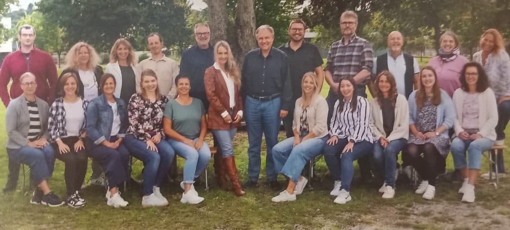 Eine Gruppe von 24 Erwachsenen, Männer und Frauen, stellt sich in zwei Reihen im Freien für ein Gruppenfoto auf. Sie sind leger gekleidet, die meisten lächeln, und im Hintergrund sind Bäume zu sehen.